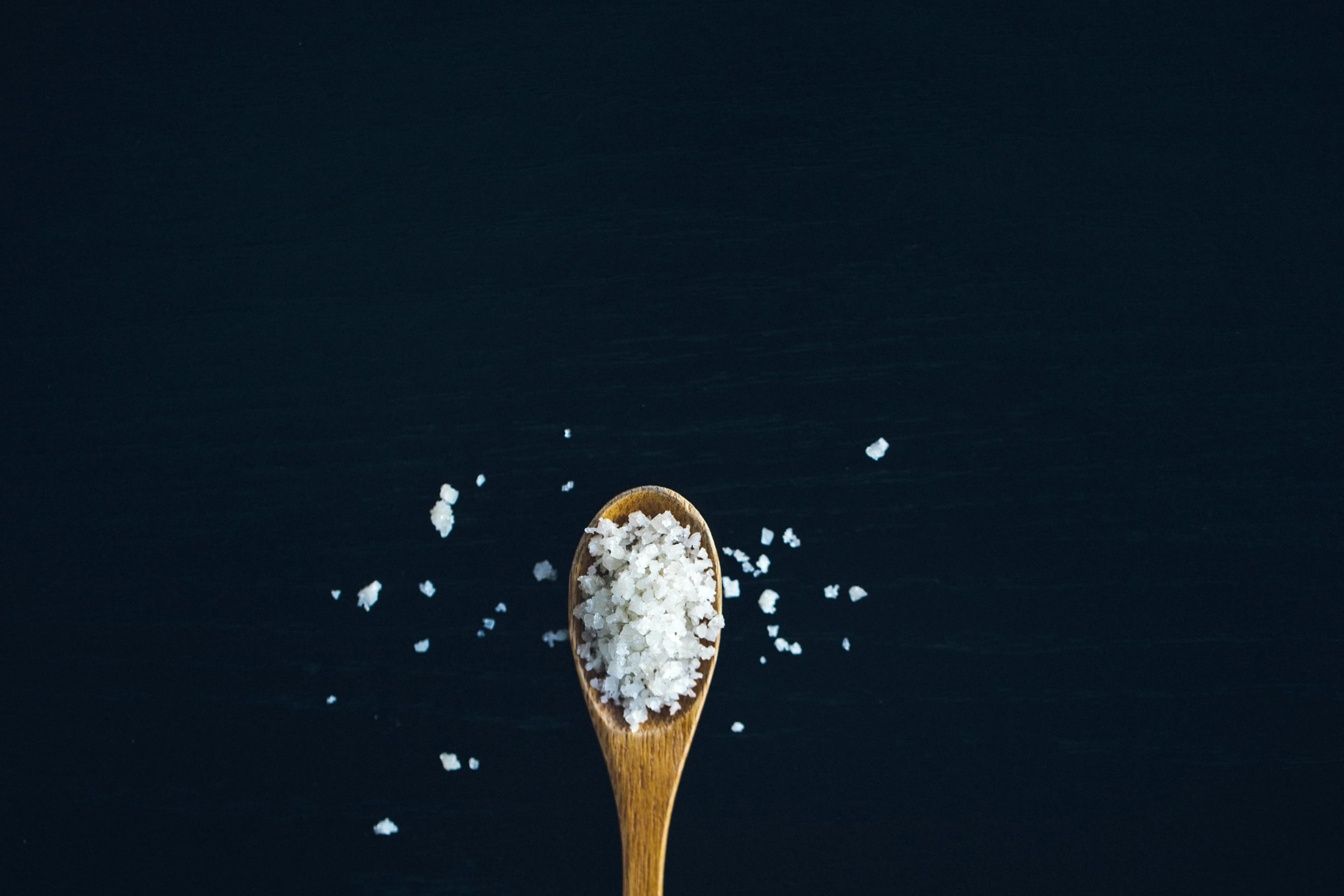 Дървена лъжица с крупна морска сол Fleur de Sel е поставена върху тъмен, черен фон, като съдържащата се сол е частично разпръсната около лъжицата.